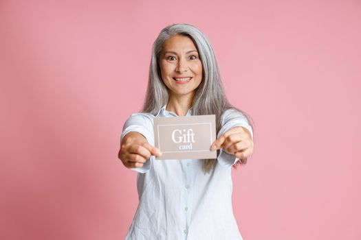 Attractive happy grey haired Asian lady holds gift card standing on pink background in studio. Shopping certificate