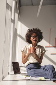 Smiling young female worker sitting at the desk and holding cup with hot drink in the office