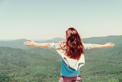 Unrecognizable happy explorer young woman standing with raised arms high in summer mountains on top.