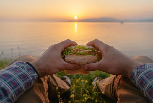 Young man holding fresh tasty burger sandwich and relaxing by the sea at summer sunset outdoor, point of view.