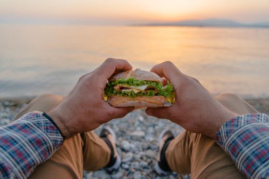 Young man holding fresh tasty burger and resting by the sea at summer sunset, point of view.