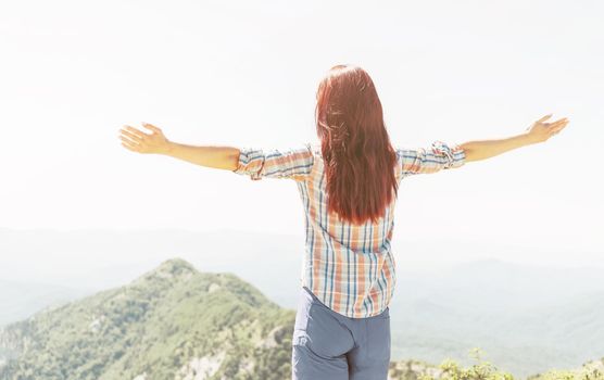 Rear view of freedom explorer young woman standing with raised arms in summer mountains.