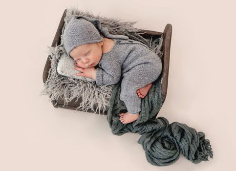 Newborn baby boy sleeping in tiny bed wearing knitted costume and holding hands under cheeks. Infant kid studio portrait