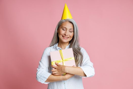 Cheerful mature Asian woman with loose grey hair and yellow party hat hugs gift box posing on pink background in studio