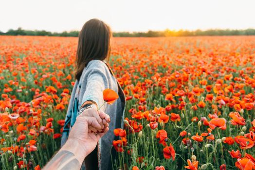 Loving couple. Man follows a brunette young woman in red poppy flower field at sunset in summer outdoor, point of view.