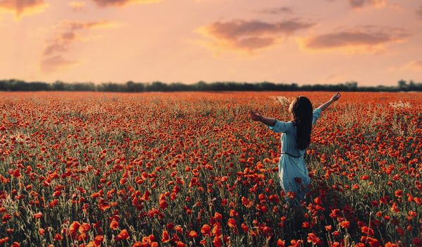 Brunette young woman with long hair relaxing with raised arms in poppy flower meadow at sunset in summer.