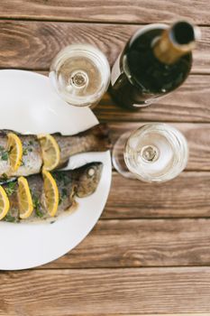 Baked fish dish on a plate and two glasses of white wine for couple on a wooden table, top view.