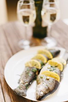 Baked fish with lemons and greenery on a plate and two glasses of white wine in the kitchen, nobody.