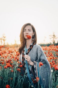 Attractive brunette young woman with long hair standing with one red poppy in flower field in summer outdoor.