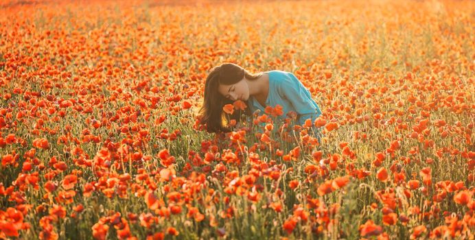 Attractive brunette young woman relaxing in flower field and sniffing a poppy in summer.