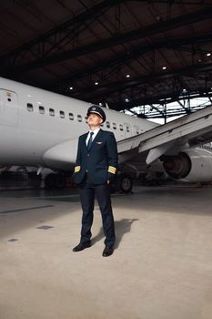 Full length shot of confident pilot in uniform looking away, ready for flight, standing in front of big passenger airplane in airport hangar. Aircraft, occupation, transportation concept