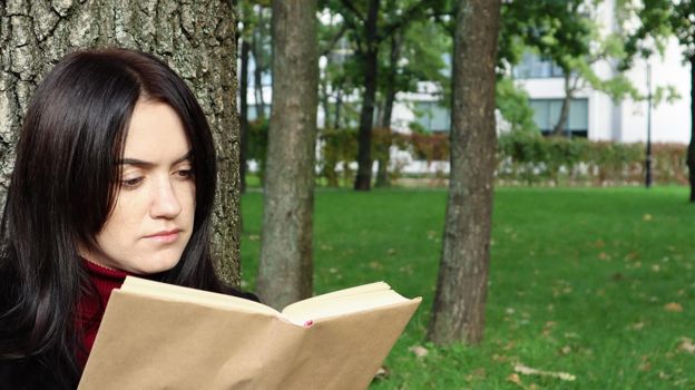 Portrait of a beautiful young woman who is sitting under a tree and reading her favorite book in a city park on green grass on a pleasant sunny day. Vacation, education and study concept