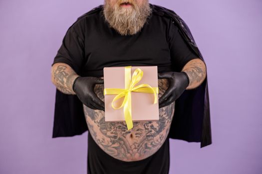 Mature bearded plus size man wearing hero suit with cape on purple background in studio closeup, focus on hands with gift box