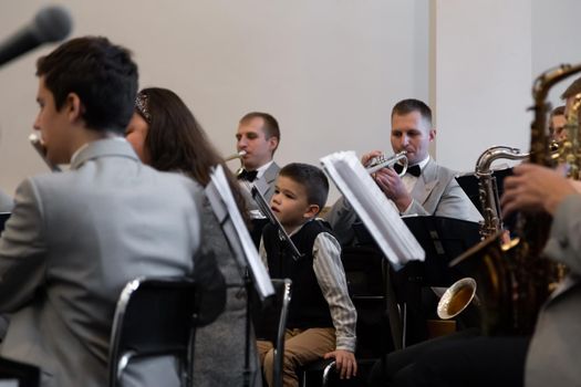 KOROSTEN - NOV, 10, 2019: A little boy sits in an orchestra near the musicians play different musical instruments in gray suits.