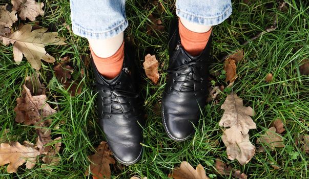 Female legs in black boots on green grass with yellow fallen autumn leaves in the park. Autumn concept. Womens shoes in autumn foliage.