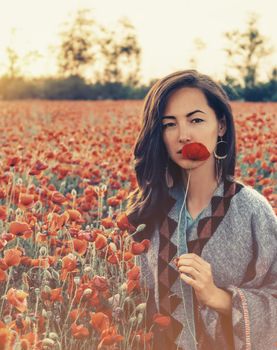 Beauty outdoor portrait of brunette young woman with red poppy near her face in flower meadow, looking at camera.