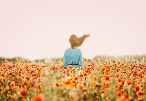 Unrecognizable brunette woman with fly-away hair standing in red poppy flower meadow in windy weather in summer, rear view.