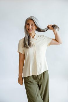 Positive Asian lady in blouse holds lock of healthy silver hair standing on light grey background in studio. Mature beauty lifestyle