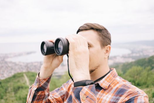 Traveler explorer young man looking through binoculars on background of sea bay in summer mountains.