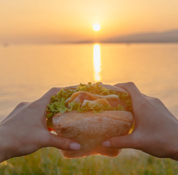 Female hands holding fresh burger in front of sea at summer sunset, point of view.