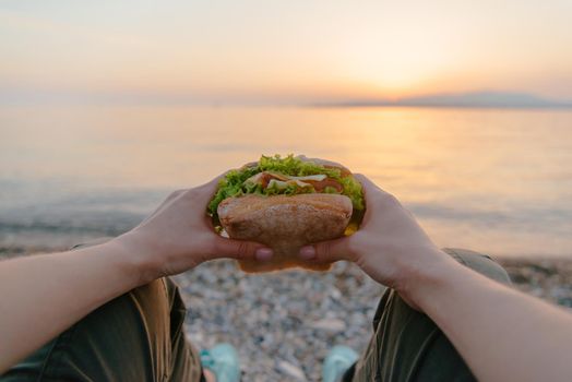 Young woman holding fresh tasty fast food burger sandwich and resting by the sea at summer sunset, point of view.