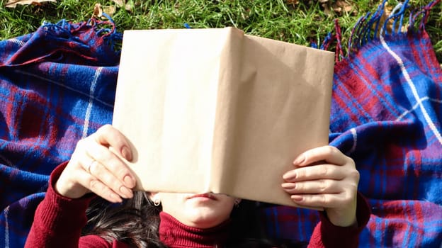 Top view of a young woman lying and reading her favorite book on a woolen plaid blanket in a park on a green grass on a pleasant sunny day. The concept of recreation, education and study