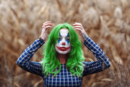 Portrait of a greenhaired woman with joker makeup on a orange leaves reeds background.