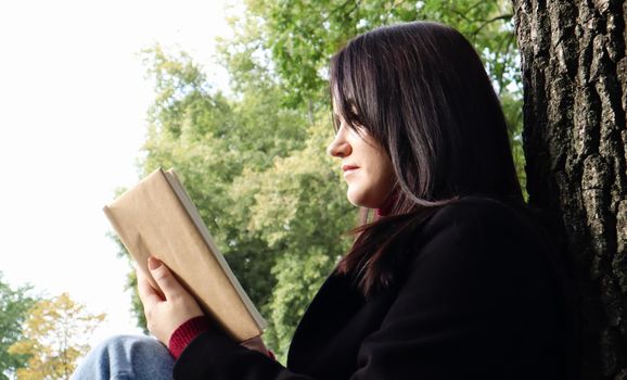 Portrait of a beautiful young woman who is sitting under a tree and reading her favorite book in a city park on green grass on a pleasant sunny day. Vacation, education and study concept