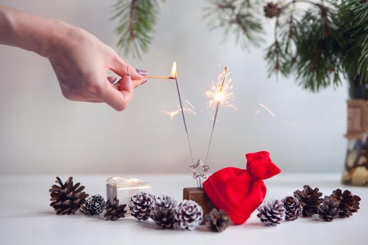 woman hand lighting festive sparkles near pine cones, christmas tree and red christmas hat. New year party burning sparkler closeup on white background. fireworks, shining fire flame. Christmas light.