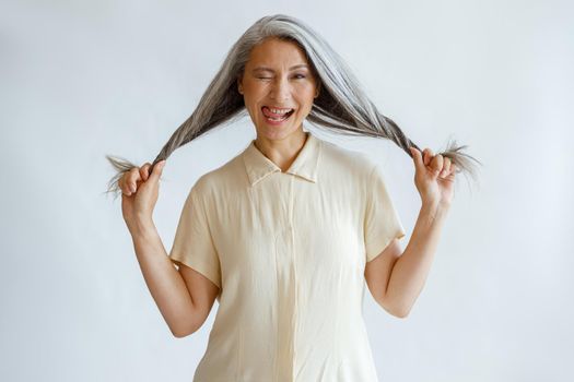 Funny middle aged Asian lady grimaces and holds long silver hair standing on light grey background in studio. Mature beauty lifestyle