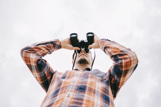 Bottom view of young man in plaid shirt watching with binoculars and exploring surroundings on white background of sky. Travel concept.