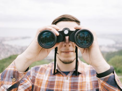 Explorer hiker young man in plaid shirt looking through binoculars on background of summer mountains and sea bay outdoor, front view. Travel concept.