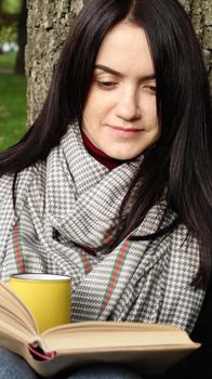 A woman sits under a tree and reads her favorite book while drinking coffee or tea from a yellow cup in a city park on green grass on a pleasant sunny day. Vacation, education and study concept