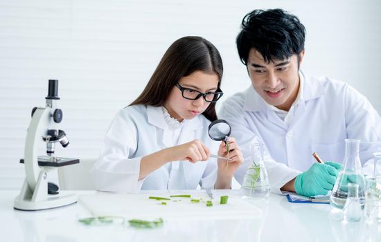 Little girl as young scientist use magnifying glass to analyze plant tissue and discuss with her teacher who sit near and consult the experiment also record the data in laboratory or classroom.