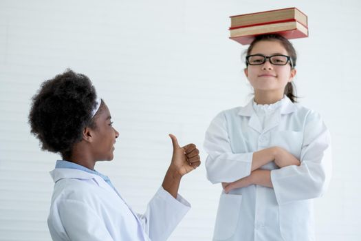 Young African American scientist girl shows thumbs up to her Caucasian friend who put stack of books on her head in laboratory or classroom.