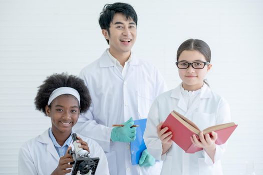 Young African American and Caucasian scientist girls with their Asian teacher stand in laboratory or classroom and look to camera with smiling during study or do some experiment.