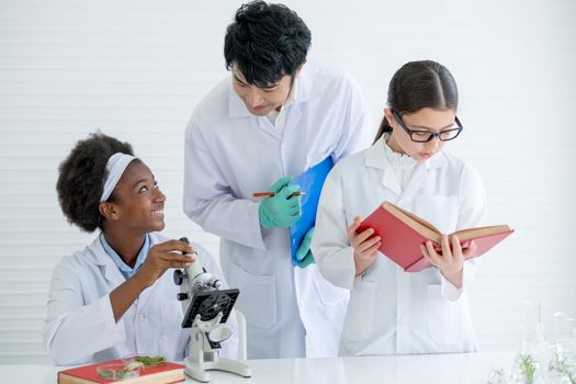 Asian teacher help and guide to African American young scientist child girl to analysis using microscope in laboratory or classroom while other student also read textbooks.