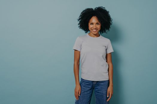 Delighted young african female isolated over blue background with pleasant smile, wearing casual white tshirt and jeans, being in good mood and demonstrating positiveness. Happy mixed race woman