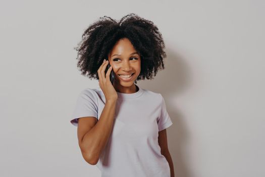 Studio portrait of cheerful dark skinned woman enjoying conversation on mobile phone, holding smartphone at smiling, hearing good news from friend while standing isolated over white background