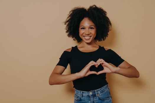 Close up photo of beautiful amazing dark skinned lady showing with hands sweetheart symbol, love message gesture, wears casual black t-shirt isolated over dark beige studio background with copy space