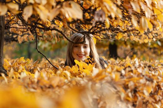 Smiling happy girl portrait with autumn leaves. Young woman among golden autumn leaves. Romantic moment in warm light. Park with yellow leaves and autumn atmosphere.