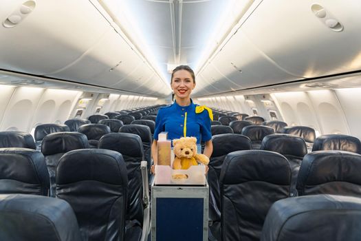 Happy pretty stewardess leading trolley cart with gifts, souvenirs through empty plane aisle