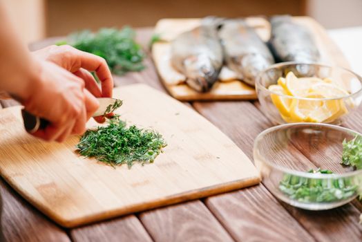 Unrecognizable woman cutting greenery on wooden board in the kitchen for fish dish on dinner, view of hands.