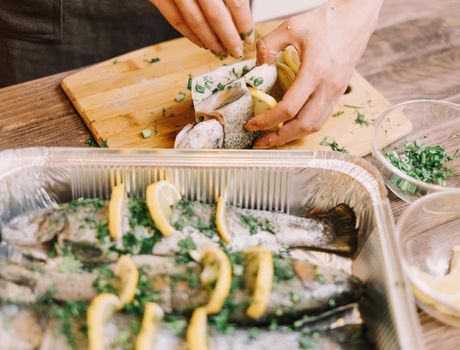 Unrecognizable woman preparing raw fish trout with lemons and greenery in the kitchen, view of hands.