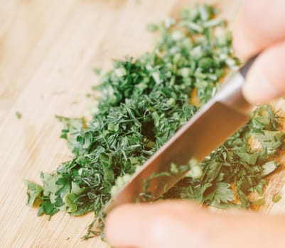 Female hands cutting parsley and dill greenery on wooden board, close-up.