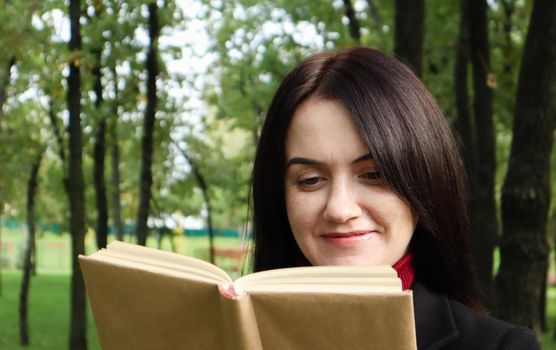 Smiling brunette woman reading a book in the park. Portrait of a happy charming Caucasian woman outdoors