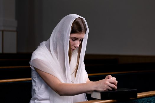 A young modest girl with a handkerchief on her head and a bible in her hands is sitting in church and praying. The concept of religion, prayer, worship.