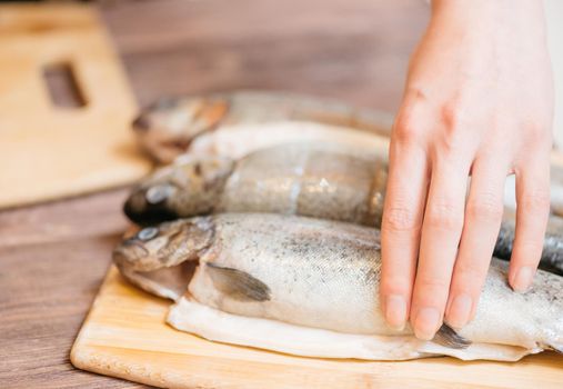 Raw fish trout on wooden board for seafood dish, woman preparing dinner in the kitchen, view of hands.