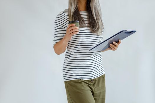 Slim middle aged woman with silver hair in casual clothes holds clipboard and cup on light grey background in studio closeup