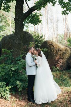 Bride and groom on wedding day, hugging, standing near a rock or a large stone.